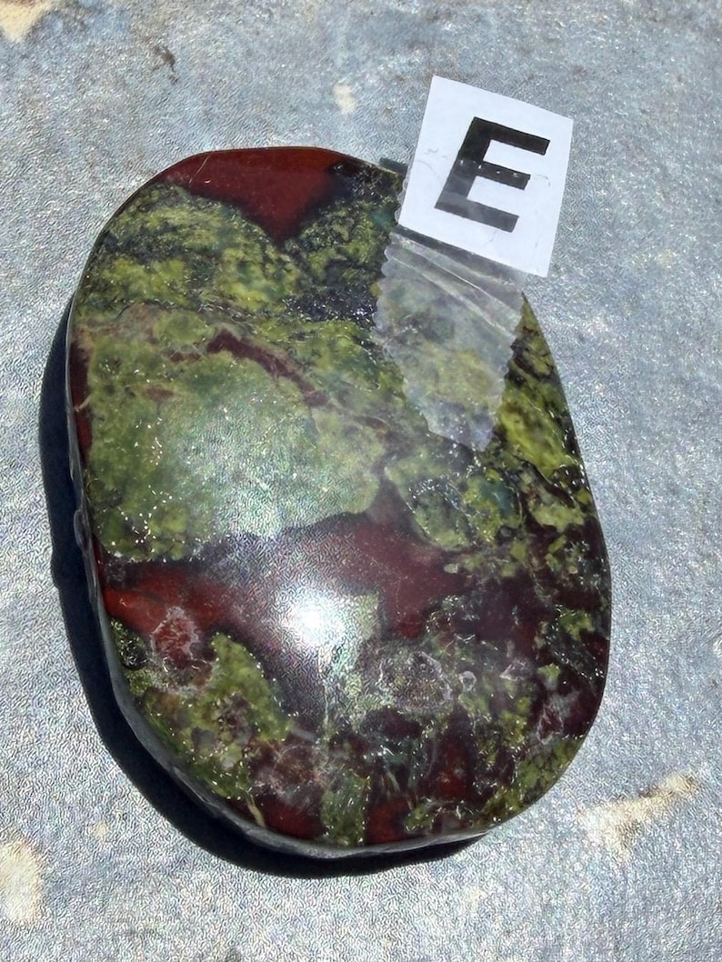 Puede incluir: Una piedra pulida de forma ovalada con una mezcla de tonos rojo oscuro y verde. La piedra tiene un acabado brillante y un aspecto natural y org&aacute;nico. Una peque&ntilde;a etiqueta blanca con la letra "E" est&aacute; adherida.