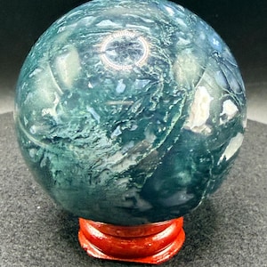 Puede incluir: Una esfera de piedra preciosa pulida, con un dise&ntilde;o arremolinado en verde y blanco, exhibida sobre un peque&ntilde;o soporte de madera rojo. La superficie pulida de la esfera refleja la luz. Un art&iacute;culo decorativo, de unos 8 cm de di&aacute;metro.
