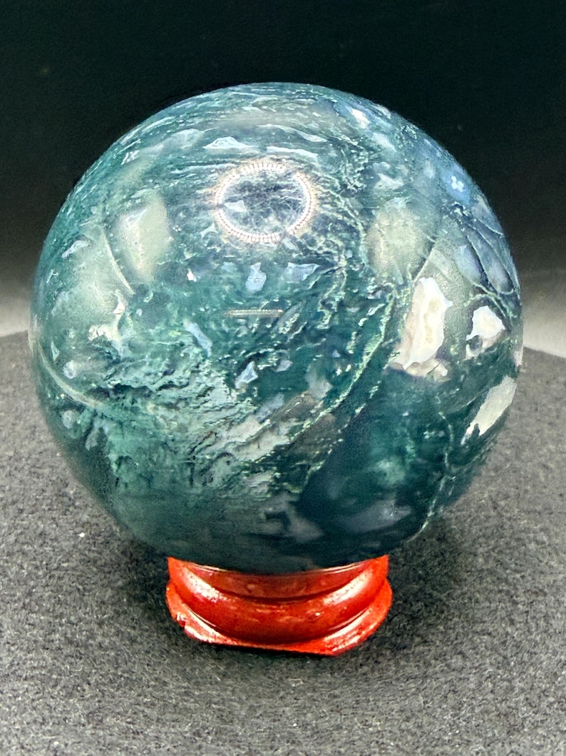 Puede incluir: Una esfera de piedra preciosa pulida, con un dise&ntilde;o arremolinado en verde y blanco, exhibida sobre un peque&ntilde;o soporte de madera rojo. La superficie pulida de la esfera refleja la luz. Un art&iacute;culo decorativo, de unos 8 cm de di&aacute;metro.