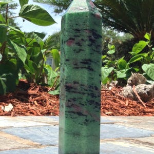 May include: A polished, hexagonal crystal tower featuring a vibrant green base color with prominent, irregular black mineral inclusions and scattered deep magenta-colored patches throughout the stone's smooth, vertical, and faceted surface.