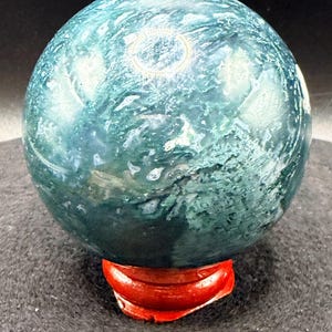 Puede incluir: Una esfera de piedra preciosa pulida, con un patr&oacute;n arremolinado de color verde azulado y blanco, colocada sobre un peque&ntilde;o soporte de madera rojo. La superficie de la esfera refleja la luz, mostrando sus texturas naturales. Ideal para decoraci&oacute;n.