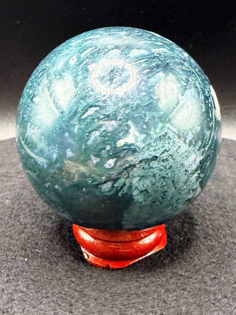 Puede incluir: Una esfera de piedra preciosa pulida, con un patr&oacute;n arremolinado de color verde azulado y blanco, colocada sobre un peque&ntilde;o soporte de madera rojo. La superficie de la esfera refleja la luz, mostrando sus texturas naturales. Ideal para decoraci&oacute;n.