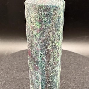 May include: A tall, rectangular crystal point with a faceted top. The stone has a mottled appearance with shades of green, purple, and white. The crystal is standing upright against a dark background.
