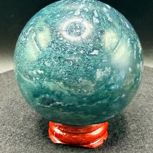Puede incluir: Una esfera de piedra preciosa pulida, con un patr&oacute;n moteado en verde azulado y blanco. Se apoya sobre un peque&ntilde;o soporte de madera rojo brillante. La superficie de la esfera refleja la luz.
