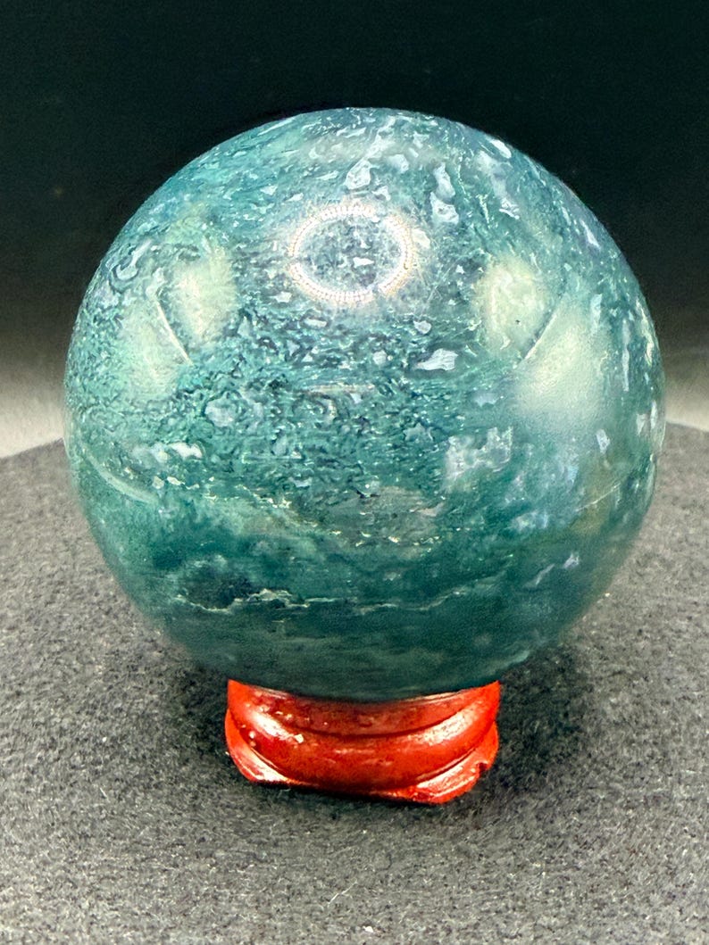 Puede incluir: Una esfera de piedra preciosa pulida, con un patr&oacute;n moteado en verde azulado y blanco. Se apoya sobre un peque&ntilde;o soporte de madera rojo brillante. La superficie de la esfera refleja la luz.