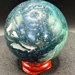 Puede incluir: Una esfera de piedra preciosa pulida, en tonos verde azulado y verde oscuro, colocada sobre un peque&ntilde;o soporte de madera rojo. El orbe tiene un aspecto veteado con patrones arremolinados, ideal para exhibir.