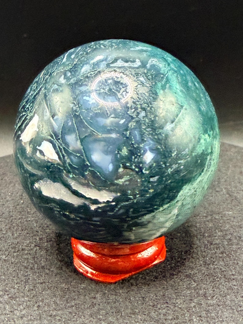 Puede incluir: Una esfera de piedra preciosa pulida, en tonos verde azulado y verde oscuro, colocada sobre un peque&ntilde;o soporte de madera rojo. El orbe tiene un aspecto veteado con patrones arremolinados, ideal para exhibir.