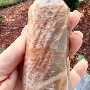 May include: A polished, rectangular crystal tower with a pointed top. The stone features a blend of peach, tan, and white hues, with natural patterns. The crystal is held in a hand, showcasing its size and texture. This item is a decorative crystal.