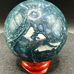 Puede incluir: Una esfera pulida de &aacute;gata musgosa, con patrones verdes y blancos. La esfera se exhibe sobre un peque&ntilde;o soporte de madera rojo. La superficie del &aacute;gata tiene un aspecto natural y texturizado, con intrincadas vetas y dise&ntilde;os.