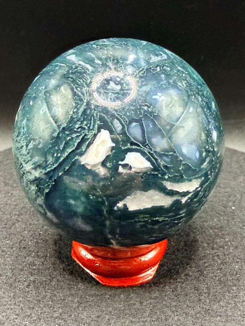 Puede incluir: Una esfera pulida de &aacute;gata musgosa, con patrones verdes y blancos. La esfera se exhibe sobre un peque&ntilde;o soporte de madera rojo. La superficie del &aacute;gata tiene un aspecto natural y texturizado, con intrincadas vetas y dise&ntilde;os.