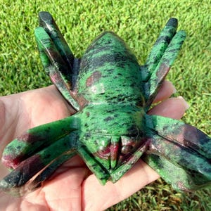 May include: A carved stone figurine in the shape of a spider, featuring a polished surface with a mottled pattern of deep forest green, black, and dark magenta hues throughout the body and the eight extended legs of the arachnid sculpture.