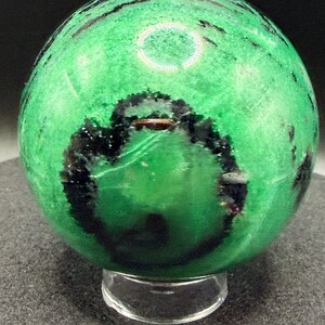 May include: A polished green sphere with black veining, resting on a clear acrylic stand. The sphere's surface has a textured appearance, and the dark veins create a striking contrast against the bright green.