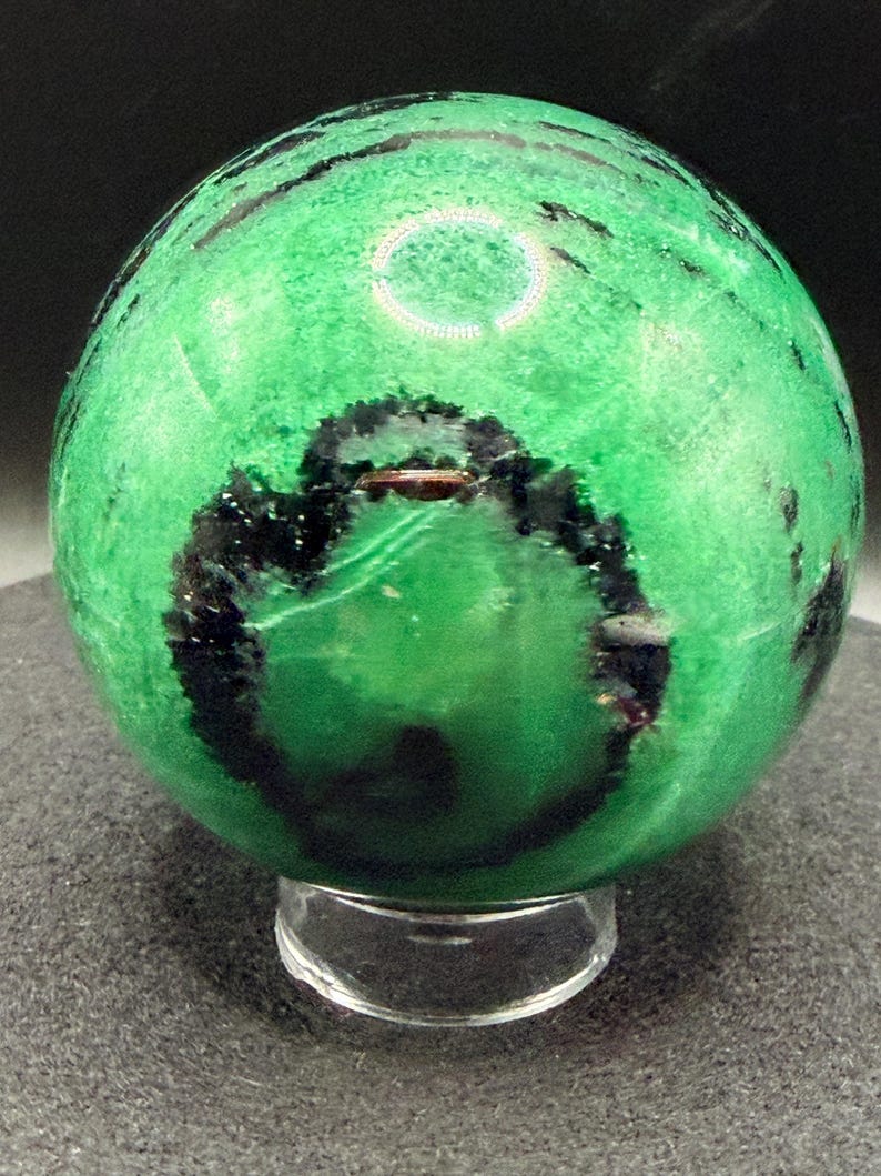 May include: A polished green sphere with black veining, resting on a clear acrylic stand. The sphere's surface has a textured appearance, and the dark veins create a striking contrast against the bright green.