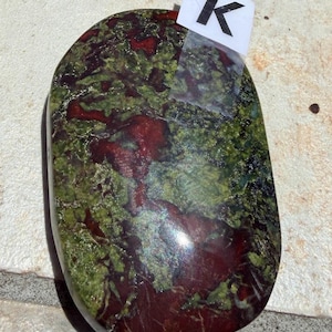 Puede incluir: Una piedra pulida de forma ovalada, con una mezcla de tonos rojo oscuro y verde. La piedra tiene un aspecto veteado, con la letra "K" en una etiqueta blanca adherida en la parte superior. Probablemente sea un objeto decorativo o una piedra preciosa.