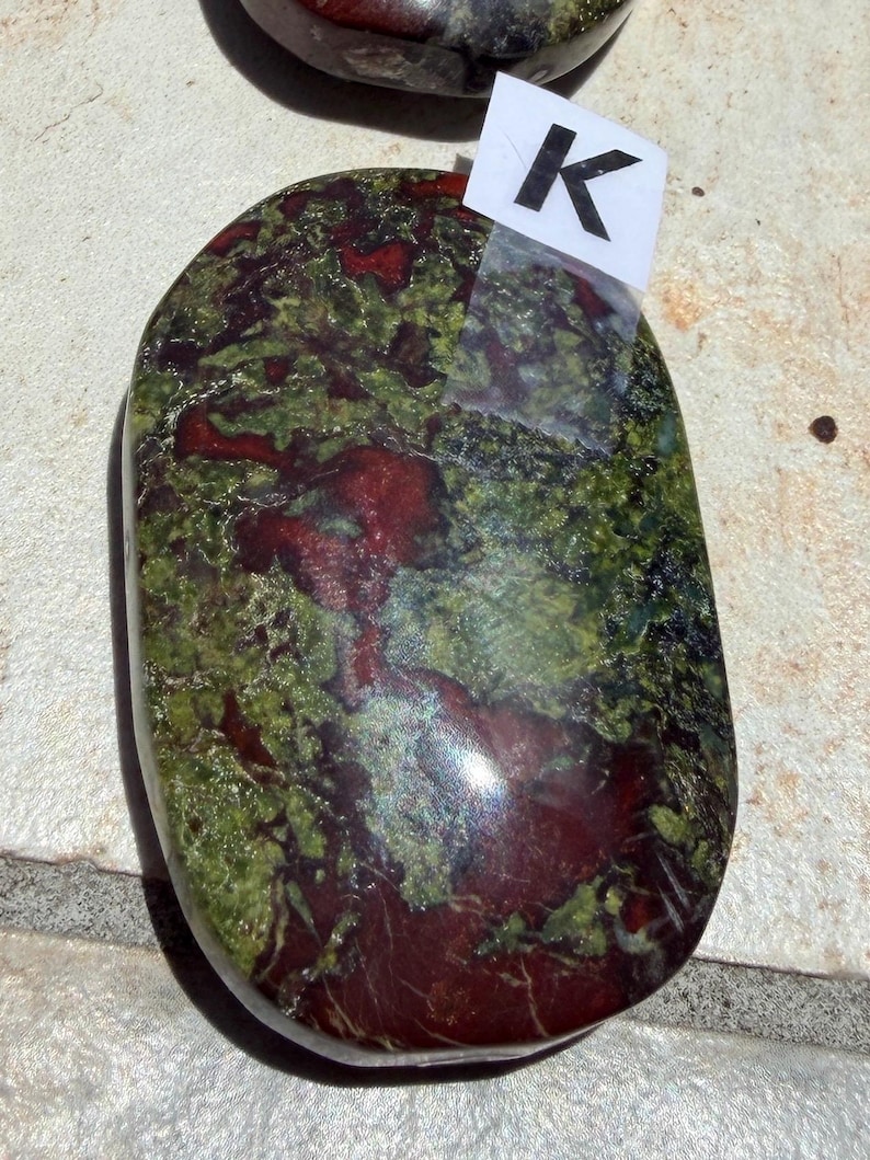 Puede incluir: Una piedra pulida de forma ovalada, con una mezcla de tonos rojo oscuro y verde. La piedra tiene un aspecto veteado, con la letra "K" en una etiqueta blanca adherida en la parte superior. Probablemente sea un objeto decorativo o una piedra preciosa.