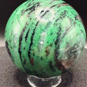 May include: A polished green zoisite sphere with black and brown inclusions, resting on a clear acrylic stand. The gemstone has a smooth, rounded shape and a vibrant color, showcasing natural patterns.