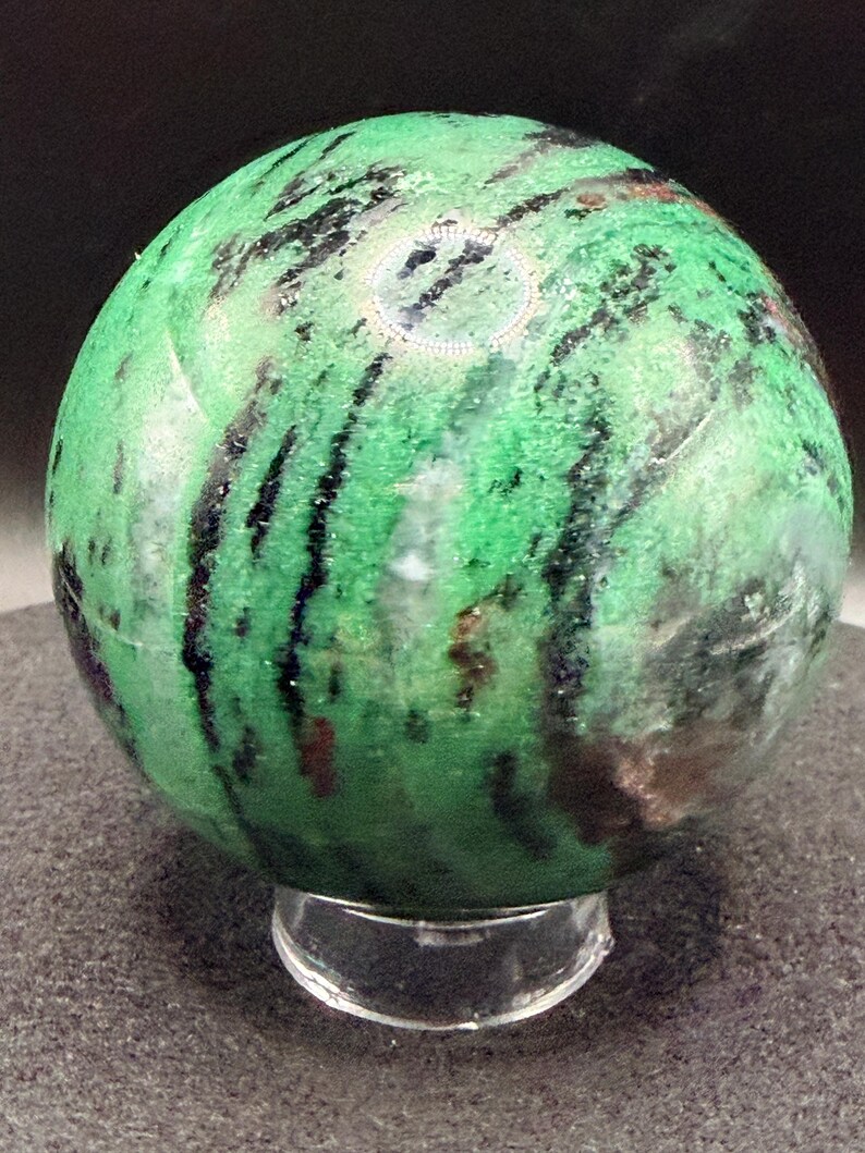 May include: A polished green zoisite sphere with black and brown inclusions, resting on a clear acrylic stand. The gemstone has a smooth, rounded shape and a vibrant color, showcasing natural patterns.