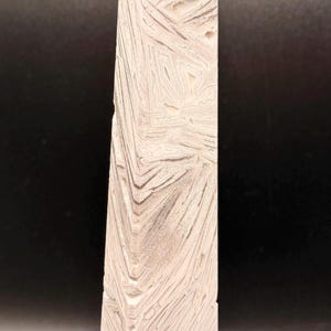 May include: A tall, rectangular, off-white stone obelisk with a textured surface. The obelisk has a pointed top and a pattern of layered, angled lines. The background is dark.