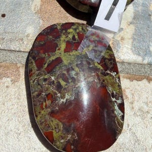 Puede incluir: Dos piedras pulidas de forma ovalada, con una mezcla de colores rojo, verde y marr&oacute;n. Las piedras tienen una superficie lisa y brillante. Una de ellas tiene una peque&ntilde;a etiqueta blanca con el n&uacute;mero 1. Estas piedras probablemente se utilizan con fines decorativos o metaf&iacute;sicos.