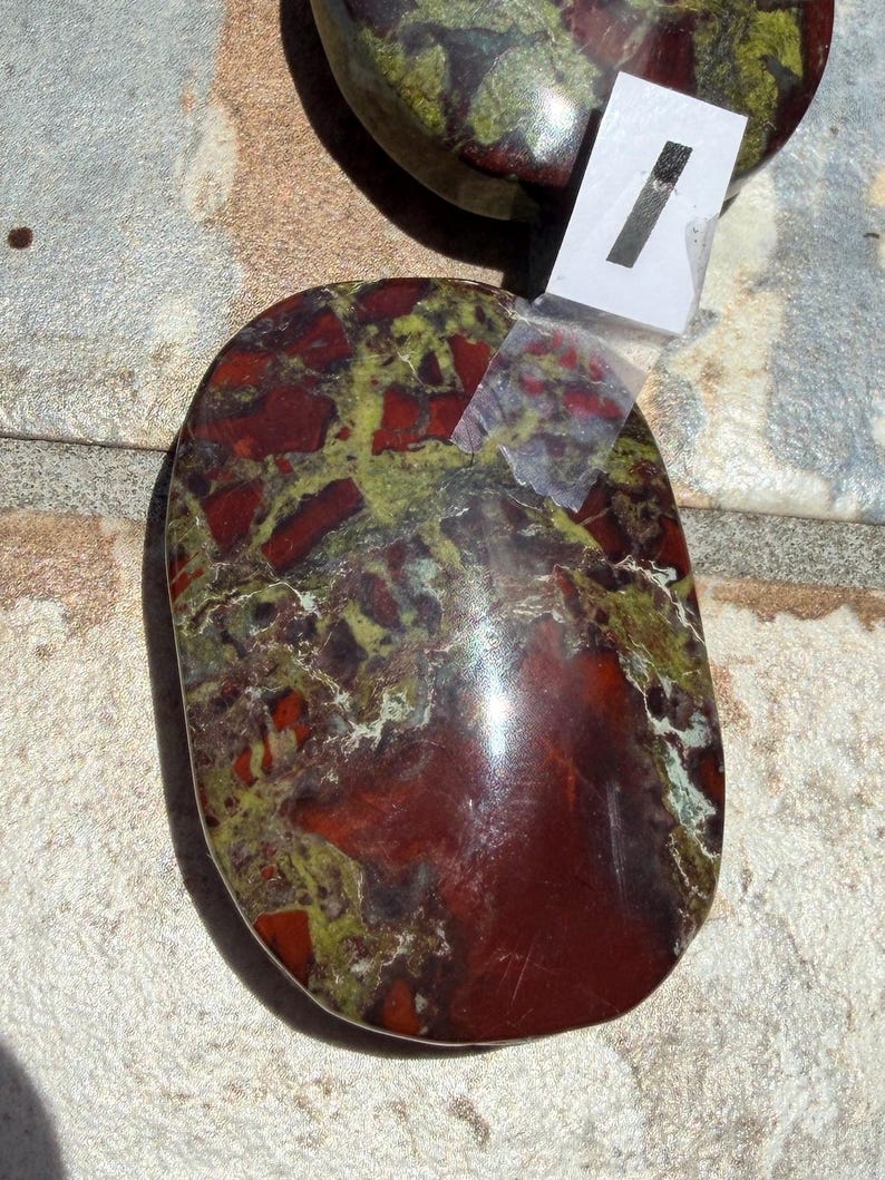 Puede incluir: Dos piedras pulidas de forma ovalada, con una mezcla de colores rojo, verde y marr&oacute;n. Las piedras tienen una superficie lisa y brillante. Una de ellas tiene una peque&ntilde;a etiqueta blanca con el n&uacute;mero 1. Estas piedras probablemente se utilizan con fines decorativos o metaf&iacute;sicos.