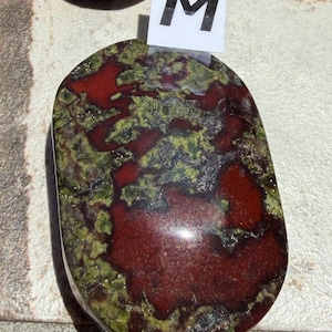 Puede incluir: Piedra pulida de forma ovalada con una mezcla de tonos rojo intenso y verde. La piedra tiene un acabado brillante y un patr&oacute;n natural y org&aacute;nico. Una peque&ntilde;a etiqueta blanca con la letra "M" est&aacute; adherida.