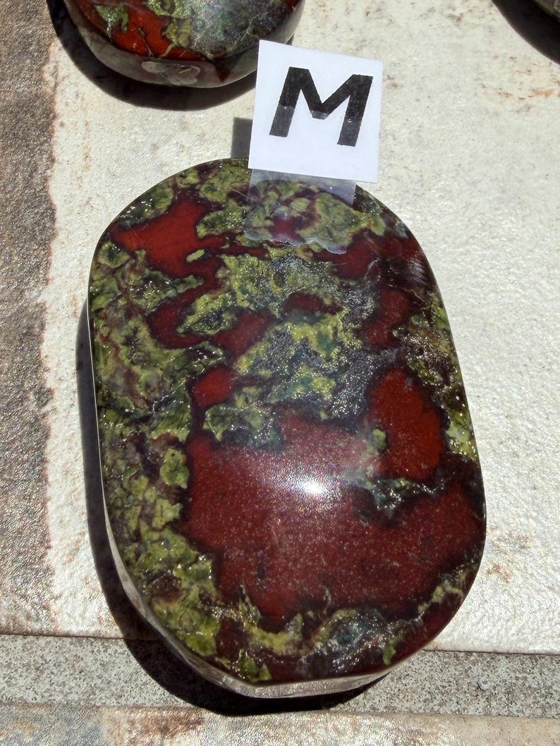 Puede incluir: Piedra pulida de forma ovalada con una mezcla de tonos rojo intenso y verde. La piedra tiene un acabado brillante y un patr&oacute;n natural y org&aacute;nico. Una peque&ntilde;a etiqueta blanca con la letra "M" est&aacute; adherida.
