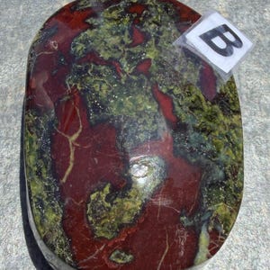 Puede incluir: Una piedra pulida de forma ovalada con una mezcla de colores rojo intenso y verde oliva. La piedra tiene un acabado brillante y un patr&oacute;n natural veteado. Una peque&ntilde;a etiqueta con la letra "B" est&aacute; adherida.