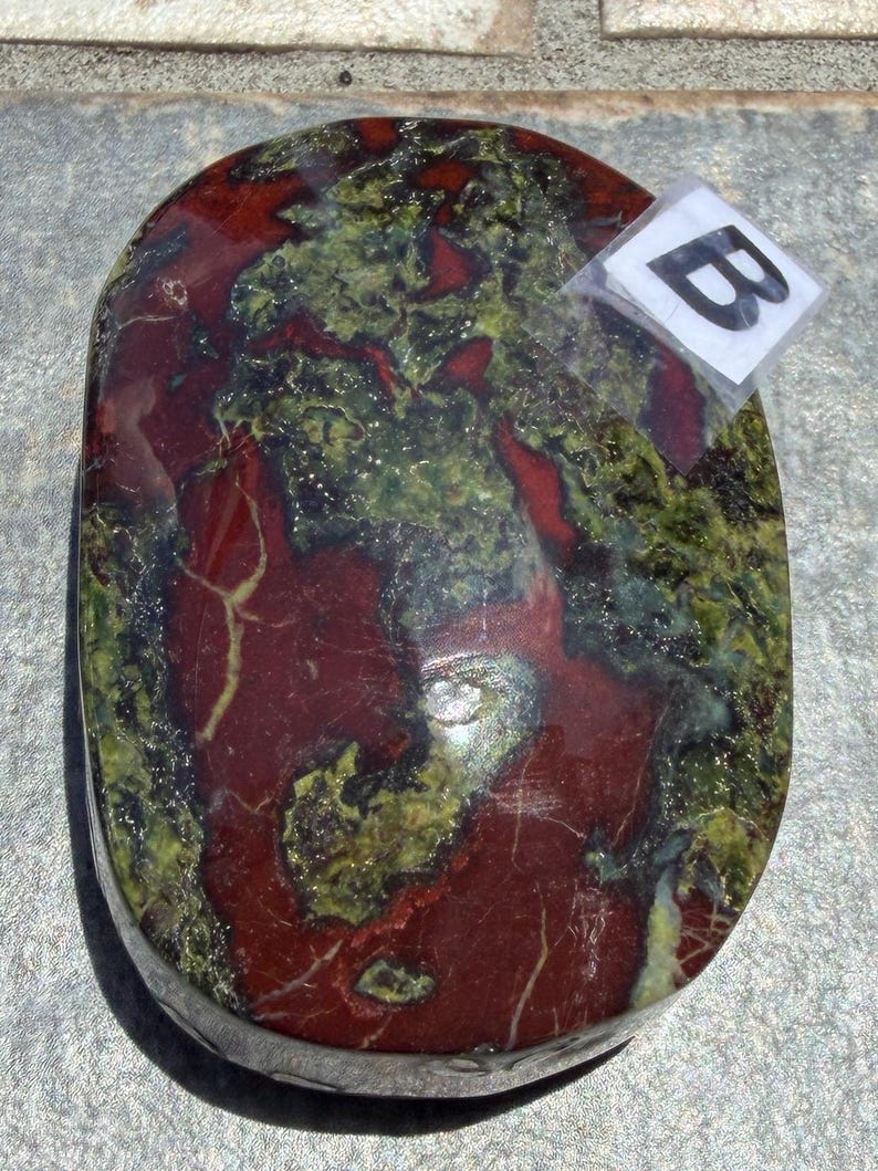 Puede incluir: Una piedra pulida de forma ovalada con una mezcla de colores rojo intenso y verde oliva. La piedra tiene un acabado brillante y un patr&oacute;n natural veteado. Una peque&ntilde;a etiqueta con la letra "B" est&aacute; adherida.