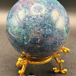 May include: A blue and multicolored sphere with a textured surface, resting on a gold-colored decorative stand. The sphere has shades of blue, green, and pink. The stand has ornate, curved legs.