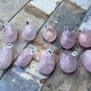 May include: An assortment of ten polished rose quartz stones, each with a unique shape and varying shades of pink. Each stone is labeled with a letter from A to J. The stones are displayed on a textured, multi-colored surface.