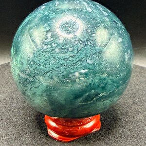 Puede incluir: Una esfera de piedra preciosa pulida, en tonos verde y verde azulado, colocada sobre un soporte de madera rojo brillante. La superficie del orbe muestra patrones y texturas intrincados. El soporte proporciona una base estable para la exhibici&oacute;n.