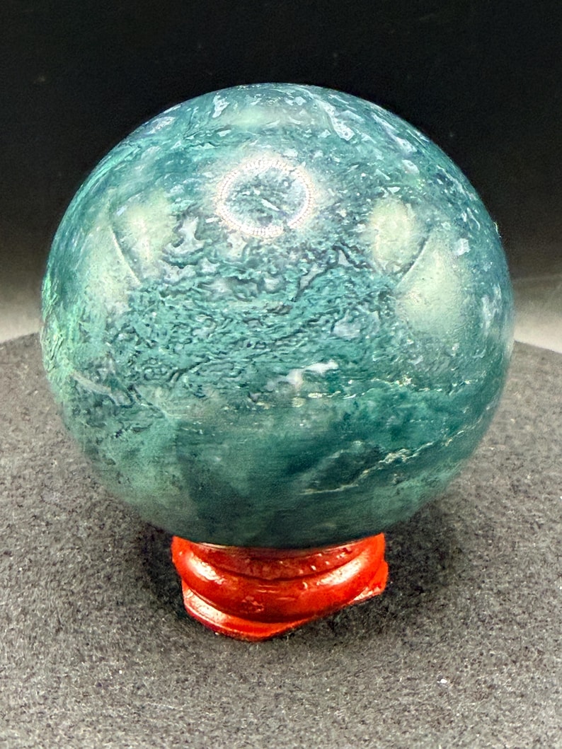 Puede incluir: Una esfera de piedra preciosa pulida, en tonos verde y verde azulado, colocada sobre un soporte de madera rojo brillante. La superficie del orbe muestra patrones y texturas intrincados. El soporte proporciona una base estable para la exhibici&oacute;n.