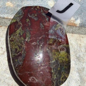 Puede incluir: Una piedra pulida de forma ovalada con una mezcla de tonos rojo oscuro, verde y negro. La piedra tiene una superficie lisa y brillante y un aspecto natural y org&aacute;nico. Una etiqueta blanca con la letra "J" est&aacute; adherida.