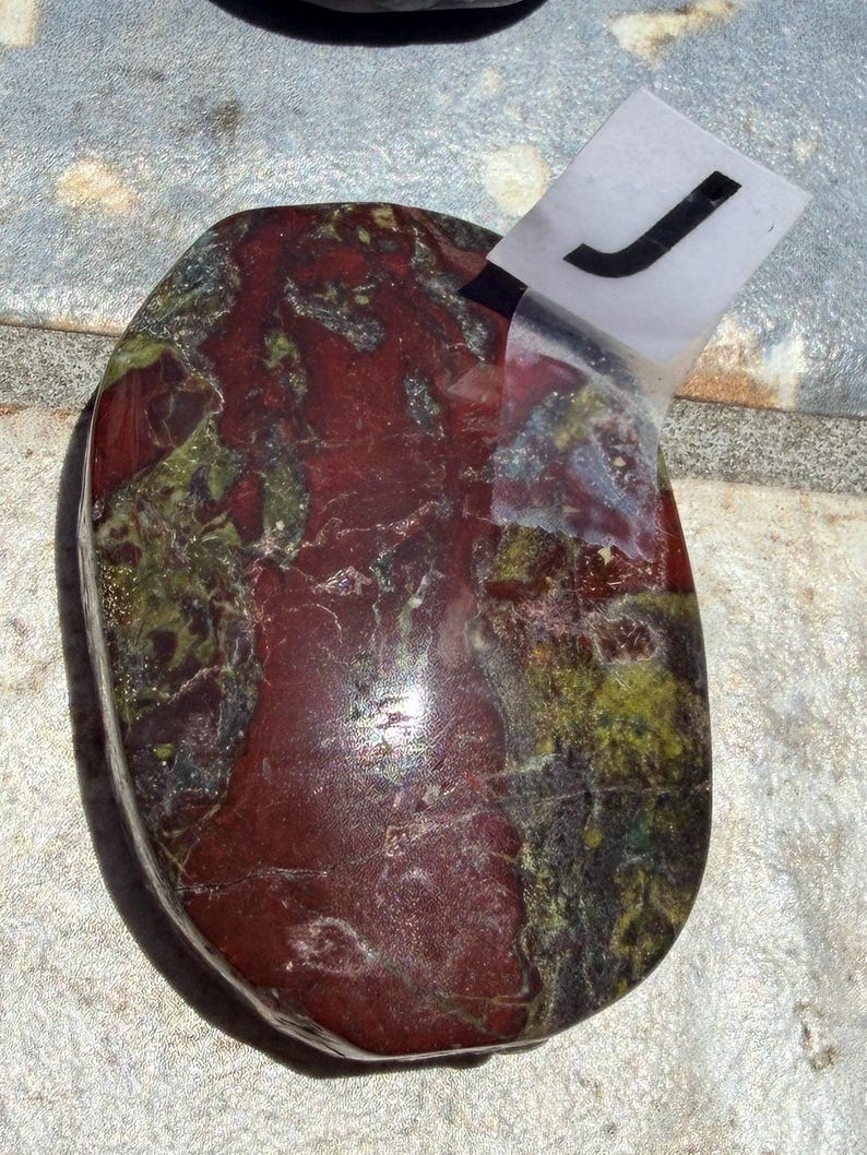 Puede incluir: Una piedra pulida de forma ovalada con una mezcla de tonos rojo oscuro, verde y negro. La piedra tiene una superficie lisa y brillante y un aspecto natural y org&aacute;nico. Una etiqueta blanca con la letra "J" est&aacute; adherida.