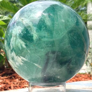 May include: A polished, spherical fluorite crystal displaying translucent shades of deep teal, emerald green, and clear sections. The stone features internal white, feathery inclusions and natural fractures throughout its rounded surface.