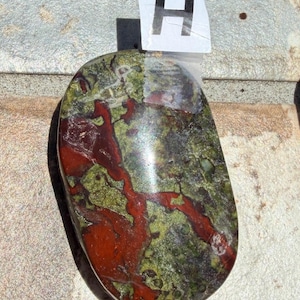 Puede incluir: Una piedra pulida de forma ovalada con una mezcla de colores. La piedra presenta una base de tonos verdes y rojos, con un patr&oacute;n de vetas rojas que la atraviesan. Una etiqueta blanca con la letra "H" est&aacute; adjunta.