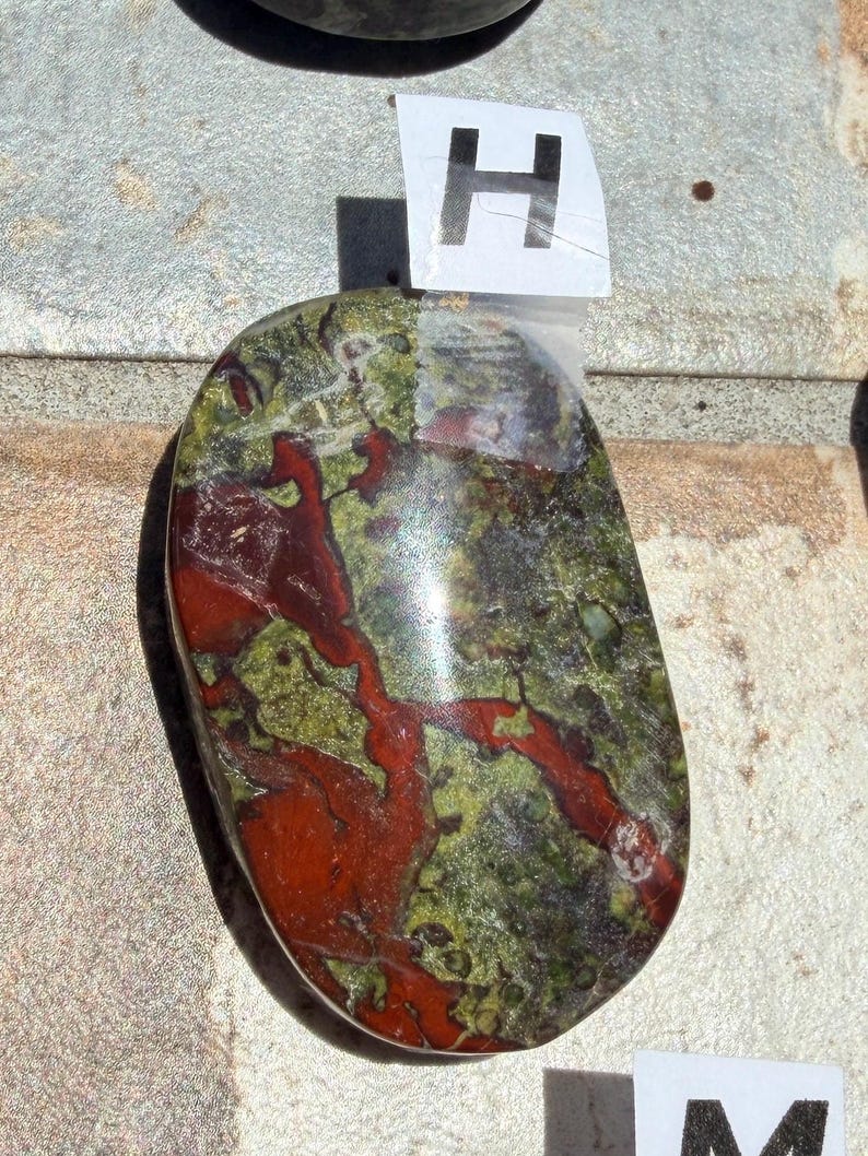 Puede incluir: Una piedra pulida de forma ovalada con una mezcla de colores. La piedra presenta una base de tonos verdes y rojos, con un patr&oacute;n de vetas rojas que la atraviesan. Una etiqueta blanca con la letra "H" est&aacute; adjunta.