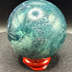 Puede incluir: Una esfera de piedra pulida, que muestra un patr&oacute;n moteado de tonos verde bosque y verde azulado intenso. La esfera se exhibe sobre un peque&ntilde;o soporte rojo brillante, creando un contraste llamativo. La superficie de la piedra tiene una textura natural y org&aacute;nica.