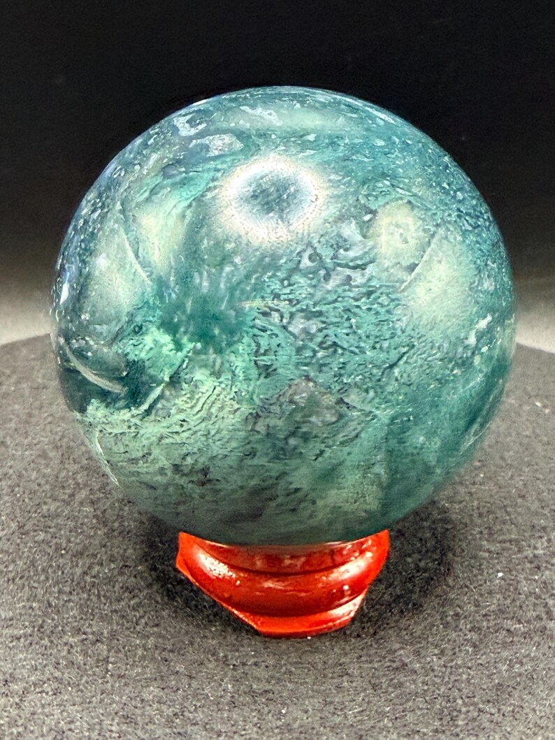 Puede incluir: Una esfera de piedra pulida, que muestra un patr&oacute;n moteado de tonos verde bosque y verde azulado intenso. La esfera se exhibe sobre un peque&ntilde;o soporte rojo brillante, creando un contraste llamativo. La superficie de la piedra tiene una textura natural y org&aacute;nica.