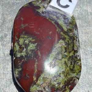 Puede incluir: Una piedra pulida de forma ovalada con una mezcla de colores. La piedra presenta una zona prominente de color rojo intenso, rodeada de patrones verdes y marrones. Una peque&ntilde;a etiqueta transparente con la letra "C" est&aacute; adjunta.