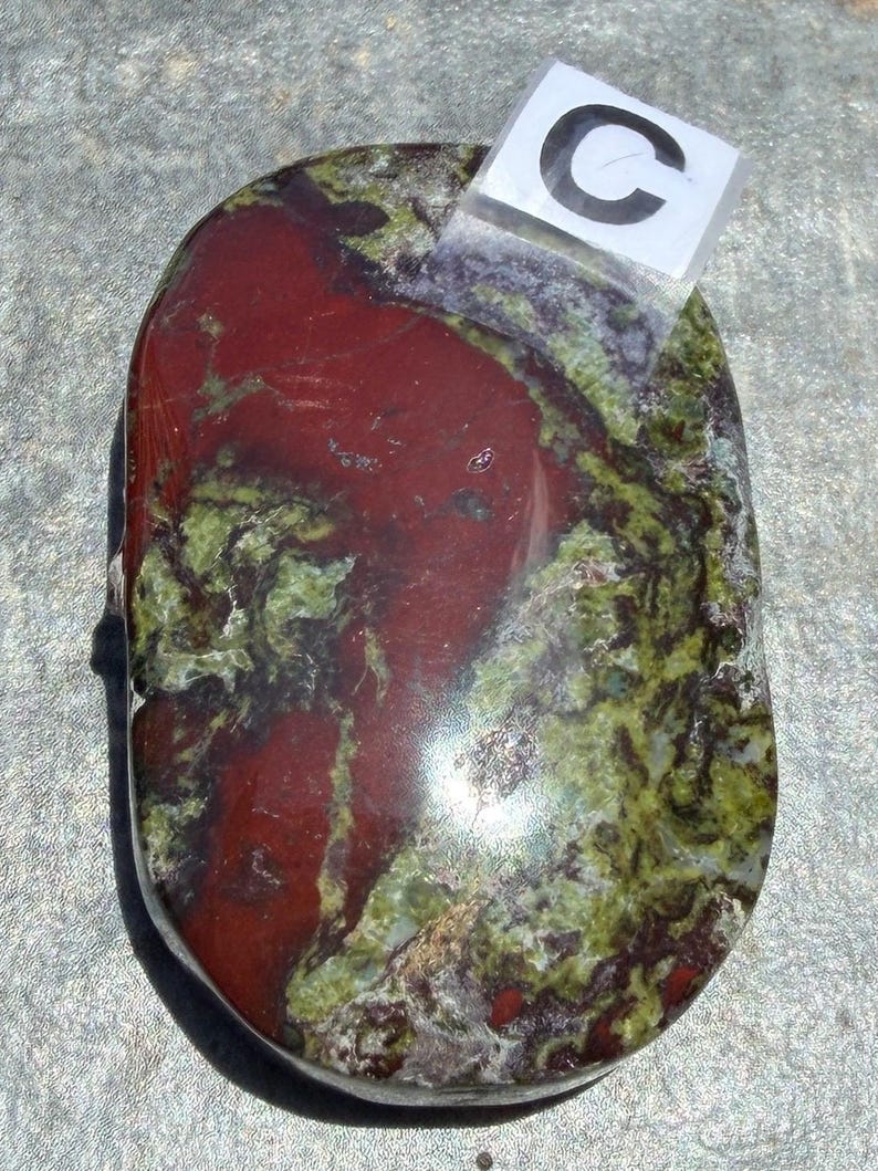 Puede incluir: Una piedra pulida de forma ovalada con una mezcla de colores. La piedra presenta una zona prominente de color rojo intenso, rodeada de patrones verdes y marrones. Una peque&ntilde;a etiqueta transparente con la letra "C" est&aacute; adjunta.