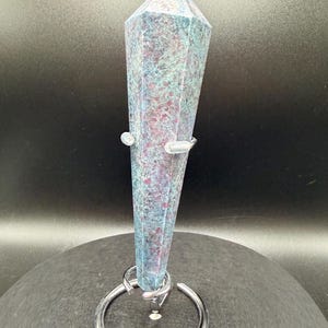 May include: A tall, faceted crystal point with a blue and purple speckled pattern. The crystal is held in place by a silver metal ring and clear plastic supports. The crystal is displayed on a dark gray surface.