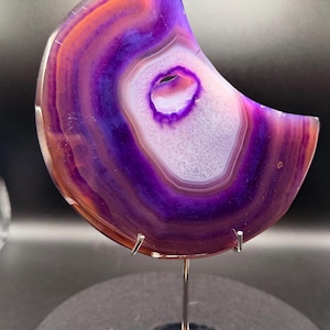 May include: A crescent-shaped agate stone with concentric bands of purple, white, and orange hues. The polished gemstone is mounted on a silver stand, showcasing its natural patterns and colors. The stone has a central, eye-shaped cavity.