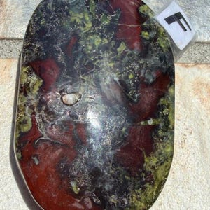 Puede incluir: Una piedra de forma ovalada con una superficie pulida. La piedra presenta una mezcla de colores, incluyendo rojo intenso, negro y verde. Una peque&ntilde;a etiqueta blanca con la letra "F" est&aacute; adherida a la piedra. La piedra tiene un acabado liso y brillante.
