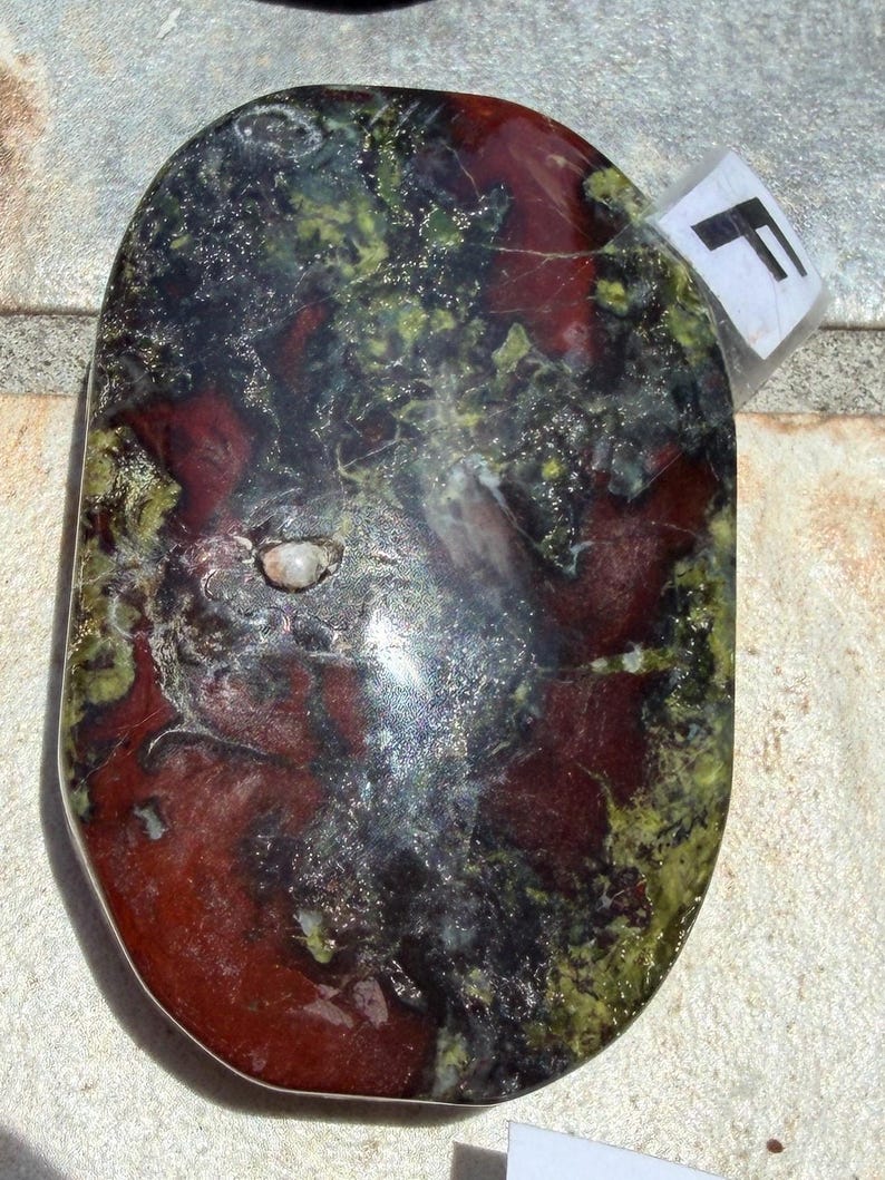 Puede incluir: Una piedra de forma ovalada con una superficie pulida. La piedra presenta una mezcla de colores, incluyendo rojo intenso, negro y verde. Una peque&ntilde;a etiqueta blanca con la letra "F" est&aacute; adherida a la piedra. La piedra tiene un acabado liso y brillante.