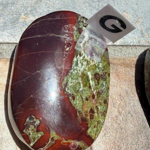 Puede incluir: Una piedra pulida de forma ovalada con una mezcla de colores. La piedra presenta una base burdeos oscuro con zonas verdes y marrones. Una peque&ntilde;a pegatina transparente con la letra "G" est&aacute; adherida.
