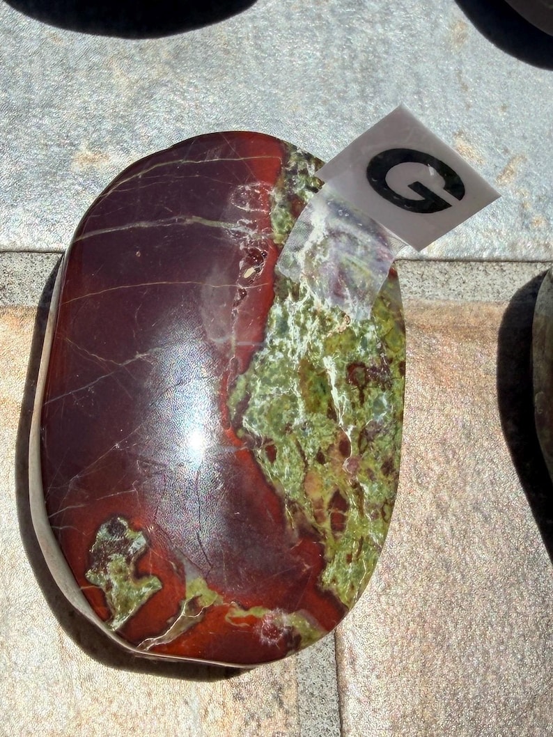 Puede incluir: Una piedra pulida de forma ovalada con una mezcla de colores. La piedra presenta una base burdeos oscuro con zonas verdes y marrones. Una peque&ntilde;a pegatina transparente con la letra "G" est&aacute; adherida.
