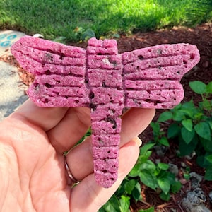 May include: A bright pink dragonfly-shaped object with a porous, textured surface. The figure features a long, segmented body and two pairs of wide, horizontal wings with horizontal grooves and small dark flecks throughout the material.