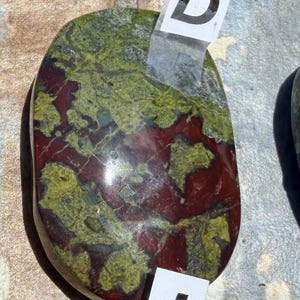 Puede incluir: Una piedra pulida, de forma ovalada, con una mezcla de colores. La piedra presenta una base roja oscura con patrones verdes y verde-amarillos. Etiquetas blancas con las letras "D" e "I" est&aacute;n adheridas. Probablemente un art&iacute;culo decorativo o una piedra preciosa.