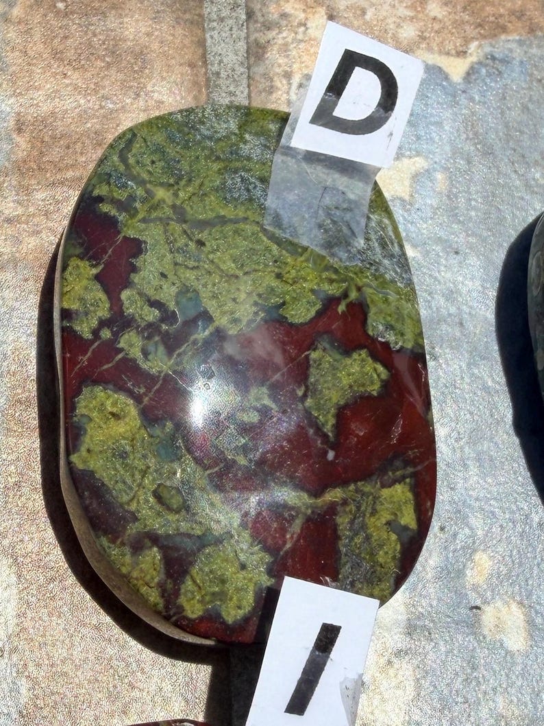 Puede incluir: Una piedra pulida, de forma ovalada, con una mezcla de colores. La piedra presenta una base roja oscura con patrones verdes y verde-amarillos. Etiquetas blancas con las letras "D" e "I" est&aacute;n adheridas. Probablemente un art&iacute;culo decorativo o una piedra preciosa.